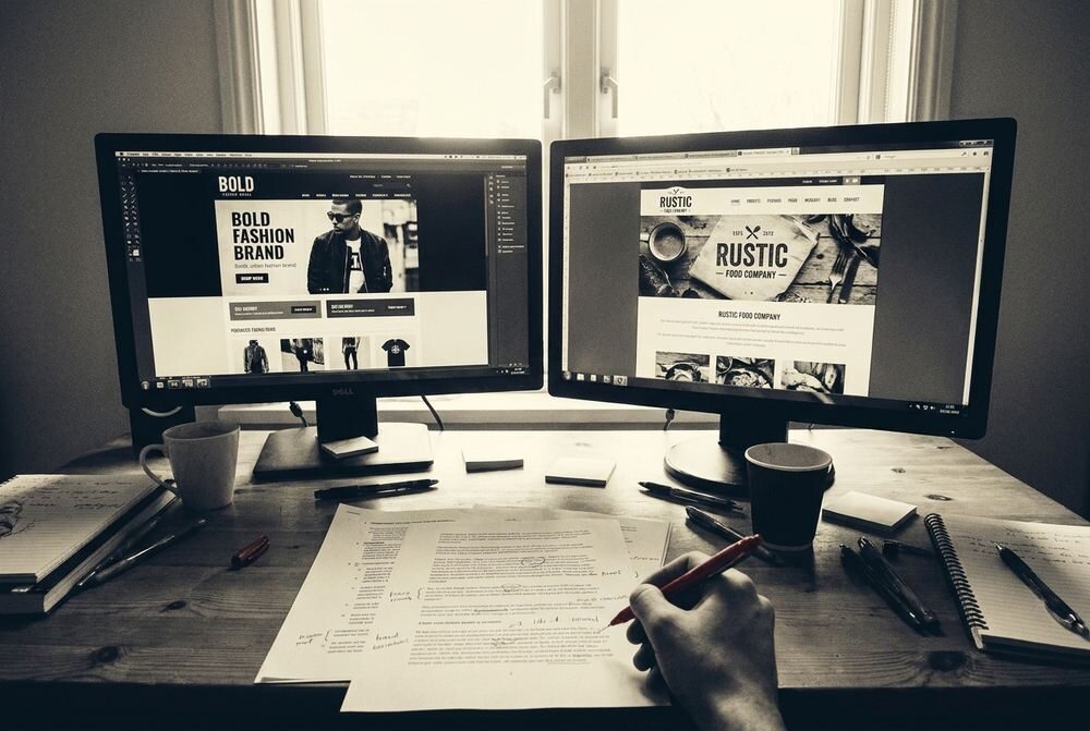 An editor's desk with two monitors showing draft layouts for different brands